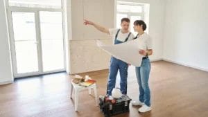 Man in overalls and woman with a blueprint discussing the best time of year to do home renovation in an empty room with painting supplies on a stool